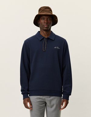 Mørkeblå langermet polo sweatshirt med krage, med en logo på brystet og ribbede mansjetter. Kombinert med en brun bøttehatt.
