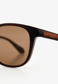 Gafas de sol con lentes marrones y montura marrón oscura. En el lateral presentan "SUPERDRY" en naranja brillante. Textura suave y diseño contemporáneo.