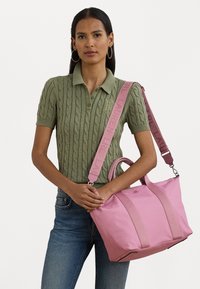 Rosa totebag med to topphåndtak og en avtakbar stropp. Har myk stoff, kontrastfargede stropper og en logo på forsiden.