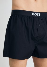 Schwarze Boxershorts aus leichtem Material, mit schlichtem Design, elastischem Bund mit "BOSS"-Logo und einer Knopfverschluss.