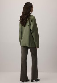 Przesadnie duży zielony sweter dziany z postarzanymi detalami, ribbowe wykończenie, w połączeniu z brązowymi spodniami dzwonami i czarnymi butami na słupku.