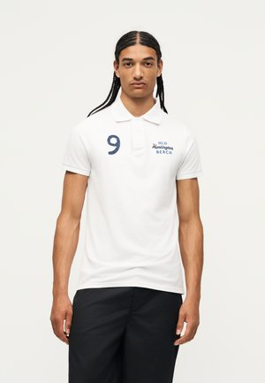 Giovane con capelli intrecciati che indossa una polo bianca con il numero 9 e la scritta blu "Huntington Beach", abbinata a pantaloni neri.