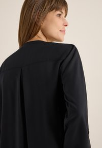 Sort bluse med en elegant, glat tekstur, en klassisk krave og en rygfold for ekstra designdetalje.