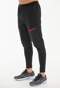Schwarze Sporthosen mit tailliertem Design, ausgestattet mit Reißverschlusstaschen und orangefarbenen Akzenten. Hergestellt aus leichtem, atmungsaktivem Material.