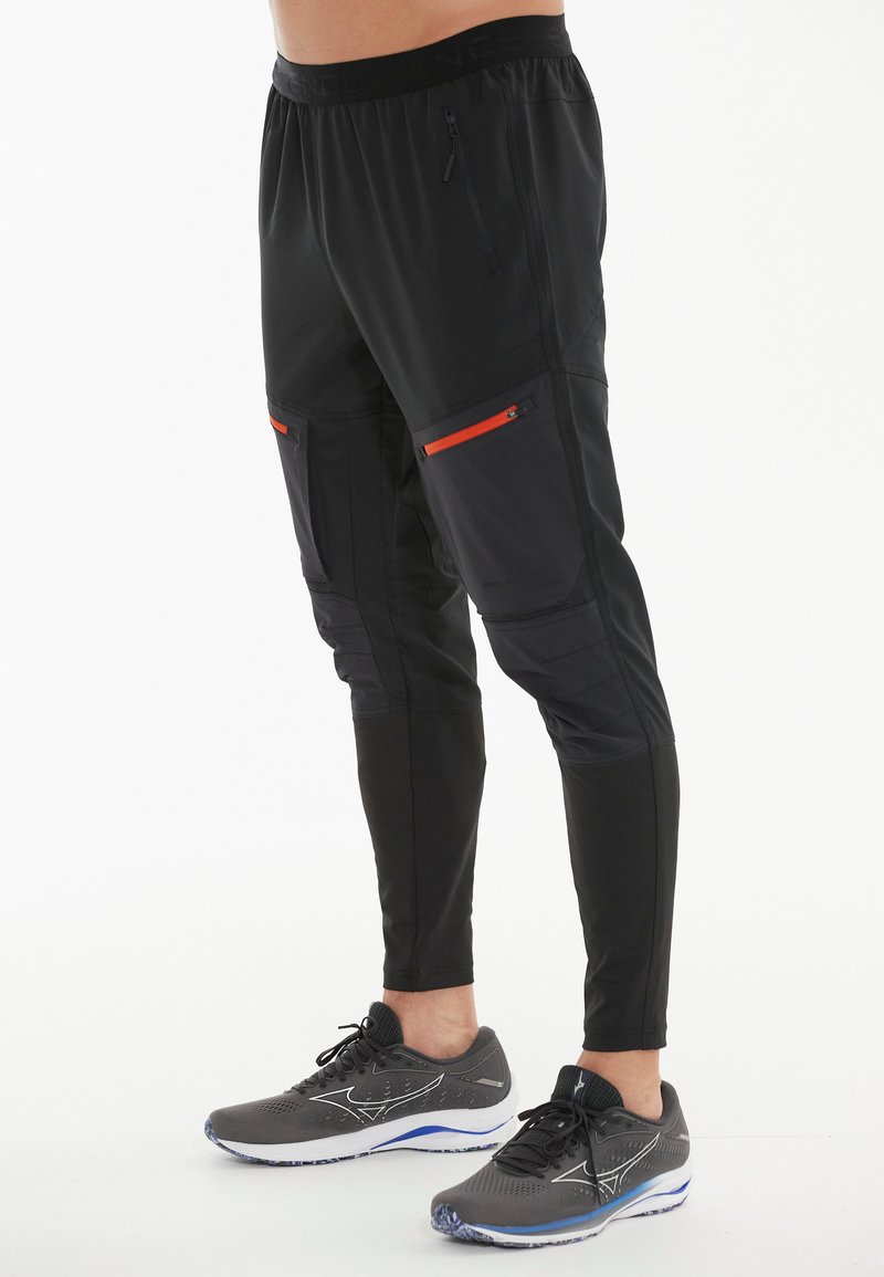 Schwarze Sporthosen mit tailliertem Design, ausgestattet mit Reißverschlusstaschen und orangefarbenen Akzenten. Hergestellt aus leichtem, atmungsaktivem Material.