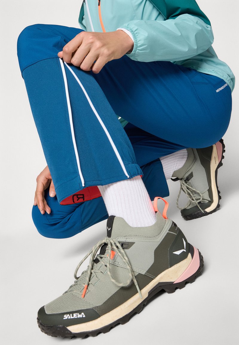 Leichte blaue Outdoor-Hosen mit weißen Seitenreißverschlüssen, kombiniert mit grauen und grünen strukturierten Sneakers mit pinken Akzenten und schwarzen Sohlen.