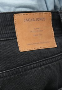 Melnas denima bikses ar brūnu ādas etiķeti, kas iešūta jostasvietā, ar iegravētu uzrakstu "JACK & JONES" un kopšanas instrukcijām tumšā drukā.
