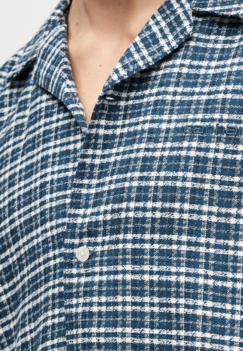 Primer plano de una camisa de cuadros azul y blanca con botones y un logotipo de marca bordado parcialmente visible en el pecho.