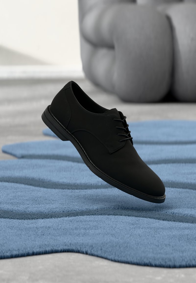 Chaussure noire à lacets flottant au-dessus d'un tapis bleu texturé, avec un canapé gris capitonné en arrière-plan.