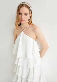 Robe blanche à couches en tissu chiffon, présentant un design plissé, un col halter avec des accents en strass et une coupe décontractée.