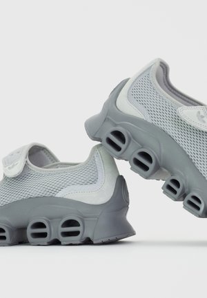 Jasnoszare buty sportowe z siateczkową cholewką i grubymi podeszwami, które mają puste, zaokrąglone prostokątne wycięcia dla amortyzacji i wentylacji.