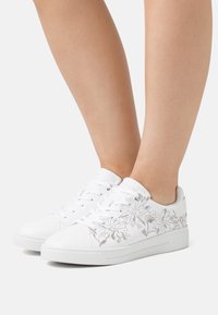 Zapatillas blancas con bordado floral plateado, punta redonda, suela plana y diseño de cordones. Textura suave con estilo minimalista.