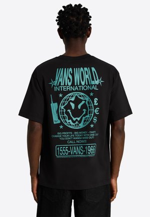 Person iført sort t-shirt med turkis grafik af forvrænget smiley ansigt, globus, telefon, valutasymboler og tekst, der promoverer "Vans World International."