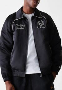 Juodas satino beisbolo švarkas su baltu apvaduku. Kairėje krūtinės pusėje yra siuvinėjamas "New York Yankees" tekstas ir logotipas. Uždarymas užtrauktuku ir elastingos rankovės.