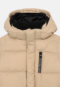 Doudoune beige avec un col haut, fermeture éclair et doublure noire. Présente un patch logo et des détails matelassés horizontaux.