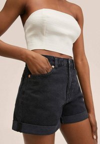 Pantalones cortos de mezclilla negra de cintura alta con puños enrollados, que cuentan con un bolsillo delantero y detalle de botón en tono cobre. La parte superior es un crop blanco sin tirantes.