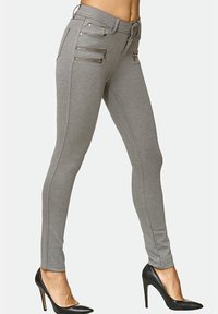 Pantaloni grigi aderenti con tasche decorative con cerniera, dettagli di cucitura sottili e una texture liscia, abbinati a tacchi alti neri.