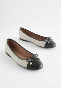 Gequiltete beige Ballerinas mit einer schwarzen, abgerundeten Zehenpartie, Schleifendetails und goldenen Metallakzenten. Glatte Innenauskleidung, flache Sohle.
