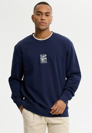 Joven con sudadera azul marino con texto gráfico blanco y pantalones beige claro, de pie frente a un fondo blanco liso.