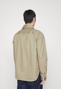 Béžová košile na knoflíky s límcem, s hladkou strukturou a švem na zadním sedle. Dlouhé rukávy s knoflíky na manžetách, stylizovaná v ležérním stylu.