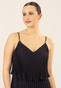 Marineblaues, plissiertes Kleid mit V-Ausschnitt und dünnen Trägern, das goldfarbene Akzente an den Schultern aufweist. Weiche, fließende Stoffstruktur.