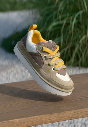 Baskets UGG beige, blanches et marron avec lacets et doublure jaunes, présentées en suspension au-dessus d'une surface en bois avec un fond de verdure.