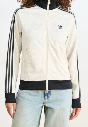 Kobieta ubrana w beżową kurtkę na zamek Adidas z czarnymi paskami na rękawach i czarnymi mankietami, zestawioną z jasnoniebieskimi dżinsami.