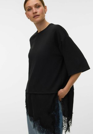 Femme portant un haut noir oversize à manches courtes avec bordure en dentelle sur un jean bleu, debout avec une main dans la poche, sur fond blanc.