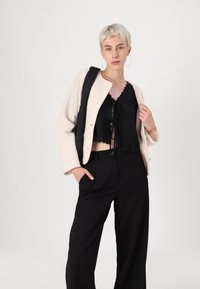Top cropped preto com renda, blazer oversized creme e calças largas pretas. Modelo segura uma mala texturizada com uma alça de ombro lisa.