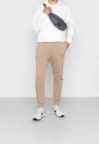 Sudadera blanca con un patrón repetitivo del swoosh de Nike, pantalones de chándal beige con un diseño similar, riñonera gris y zapatillas blancas de Nike.