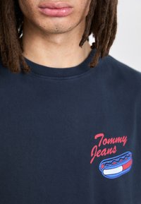 Camiseta de algodón azul marino con cuello redondo, con un bordado rojo de "Tommy Jeans" y un gráfico de perrito caliente, que presenta una textura suave y un cuello acanalado.