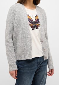 Jasnoszary dzianinowy kardigan z prążkowanym wykończeniem na dole i mankietach, z czterema różowymi guzikami. Noszony na beżowej koszulce z nadrukiem motyla.