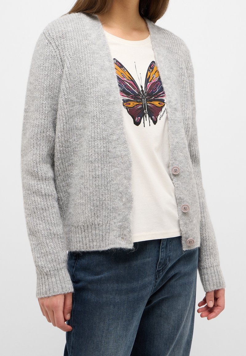 Jasnoszary dzianinowy kardigan z prążkowanym wykończeniem na dole i mankietach, z czterema różowymi guzikami. Noszony na beżowej koszulce z nadrukiem motyla.