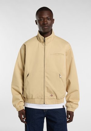 Homme portant une veste beige zippée avec poignets et ourlet élastiques, chemise blanche en dessous et jean bleu foncé, debout devant un fond uni.