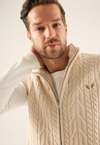 Gilet tricoté beige avec motif torsadé, fermeture zippée et détail logo subtil. Superposé à une chemise blanche texturée à manches longues.