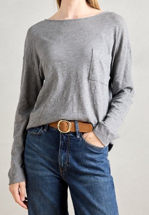 Kobieta w szarej koszulce z długim rękawem z kieszenią na piersi, niebieskich dżinsach i brązowym pasku ze złotą klamrą, z ręką w kieszeni.