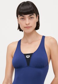 Maillot de bain bleu marine avec encolure dégagée, détail froncé à l'avant et panneau intérieur noir portant le logo "arena". Texture lisse.