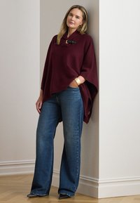 Bordeauxrød strikket poncho med høj krave og sort spænde, kombineret med blå jeans med vide ben og sorte sko med spids tå.