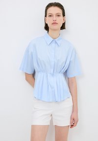 Blusa blu chiaro a maniche corte con vita aderente e fondo plissettato, dotata di colletto classico e chiusura a bottoni, abbinata a pantaloni corti bianchi.