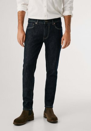 Jeans de mezclilla azul oscuro con corte ajustado, con costuras amarillas, diseño de cinco bolsillos y botas de ante marrón.