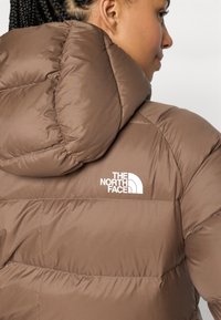 Smeđi puffer jakna s pravokutnim bijelim logom na lijevom ramenu, s prešanim uzorkom i kapuljačom.