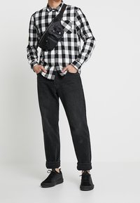 Camicia di flanella a scacchi bianco e nero, pantaloni neri arrotolati alle caviglie, sneakers nere e una borsa a tracolla nera. Look casual e stratificato.