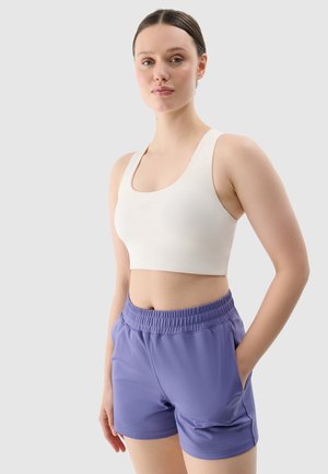 Jeune femme portant un crop top blanc sans manches et un short de sport violet, debout avec une main dans la poche sur fond gris.