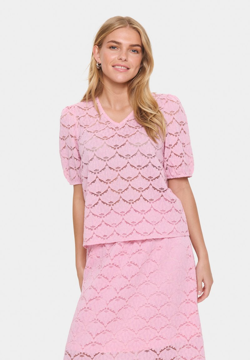 Top in pizzo rosa con maniche a sbuffo corte, scollatura a V e motivo scallop. Abbinato a una gonna coordinata, creando un look armonioso.