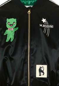 Chaqueta de satén negra con cremallera dorada, que presenta un dibujo animado de oso verde con lazo rosa, una estrella fugaz blanca con el texto "M.RODINI" y un parche de gato negro.