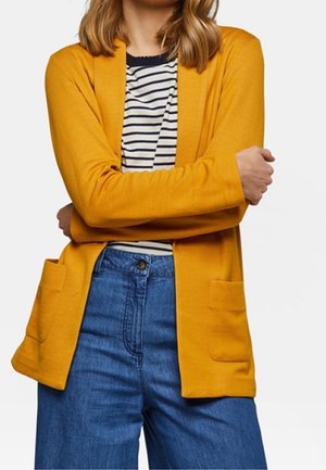 Kobieta ubrana w kardigan w kolorze musztardowym, na czarno-białej paskowanej koszuli, w połączeniu z wysokim stanem niebieskich dżinsów, z założonymi skrzyżowanymi ramionami.
