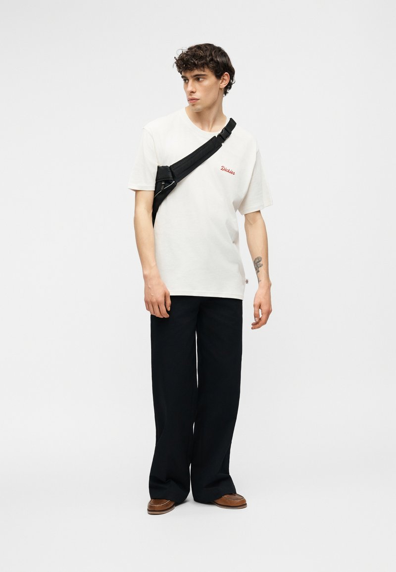 Jeune homme portant un t-shirt blanc Dickies, un pantalon large noir, des chaussures marron et un sac bandoulière noir, debout sur un fond blanc.
