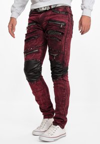 Jean bordeaux usé avec plusieurs fermetures éclair et accents de clous, comportant des patchs de genoux en cuir noir et un design slim. Baskets blanches présentées.