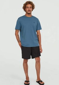 Modrá bavlnená tričko s okrúhlym výstrihom a krátkymi rukávmi, vystavené s čiernymi šortkami. Obsahuje nenápadné logo na hrudi.