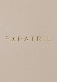 Helles rosa strukturiertes Leder mit geprägtem goldenen Text, der "EXPATRIÉ" in einer modernen Schriftart liest. Glatte Oberfläche mit subtilen Holzmaserungselementen.